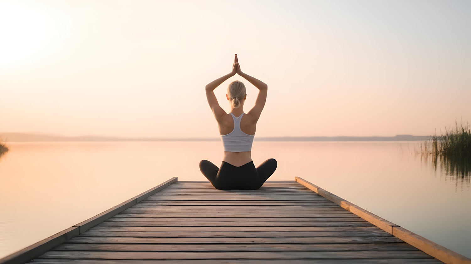 A person sitting cross-legged on a lakeside dock at sunrise with both arms raised overhead.